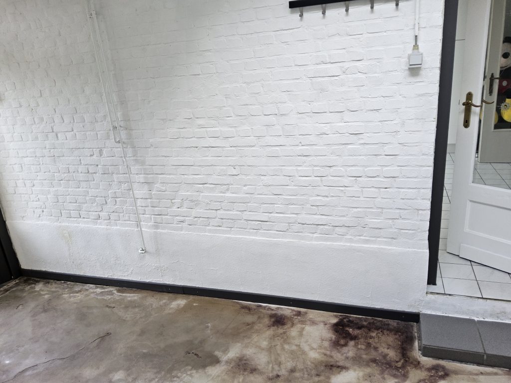 Binnenruimte met witte bakstenen muur; vochtige vloer met zichtbare bruine verkleuringen aan de rand.